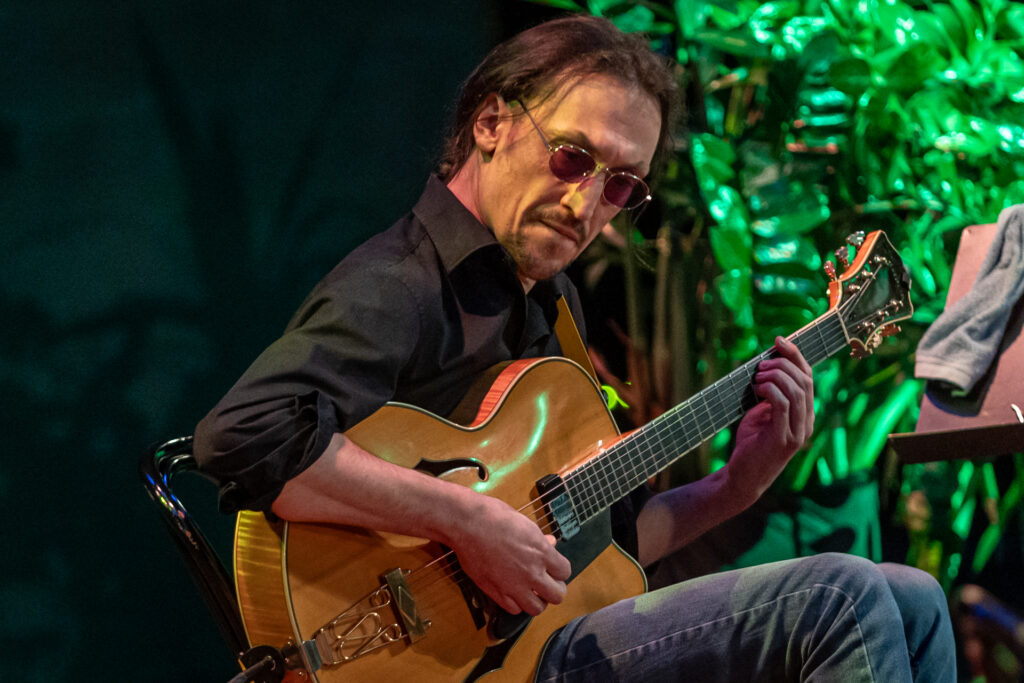 Zdjęcie. Mężczyzna gra na gitarze. Ubrany w czarną koszulę, okulary o metalowych oprawkach z brązowymi szkłami.