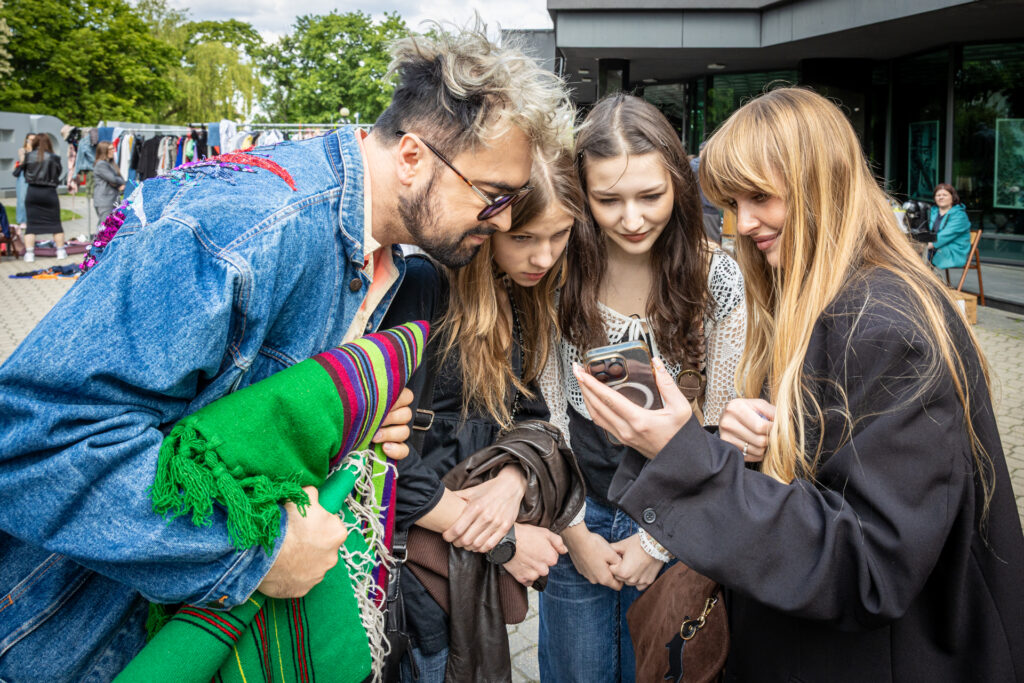 Zdjęcie. Olga Perczak-Buda pokazuje telefon młodym dziewczynom i Arturowi Wjacie. Cała trójka przygląda się ekranowi.