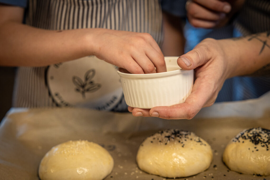 Zdjęcie. Dorosła dłoń podaje ceramiczną miseczkę. W miseczce chowa się dłoń dziecięca, która wyciąga coś drobnego. Poniżej nieupieczone bułki posypane czarnymi i białymi ziarenkami.