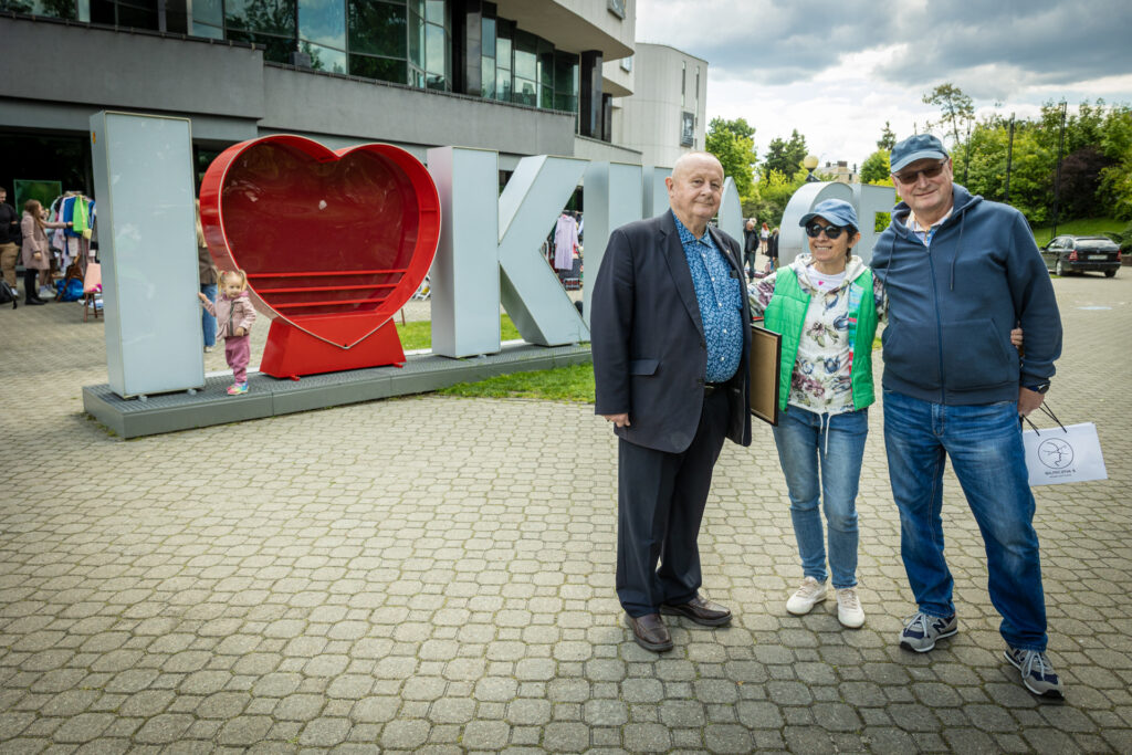 Zdjęcie. Trzy osoby: dwóch starszych panów i jedna pani po środku pozują na tle napisu I love Kielce przed budynkiem Kieleckiego Centrum Kultury.