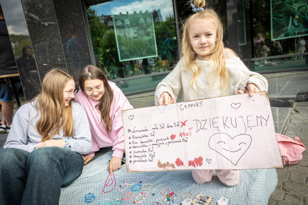 Zdjęcie. Trzy dziewczynki siedzą na kocu. Najmłodsza z nich, blondynka o długich włosach pokazuje tekturkę z cennikiem: bransoletka 5 zł, box bransoletki i pierścionki 14 zł, naszyjnik 10 zł. Po lewej strony od listy duże uśmiechnięte serce i napis: dziękujemy.
