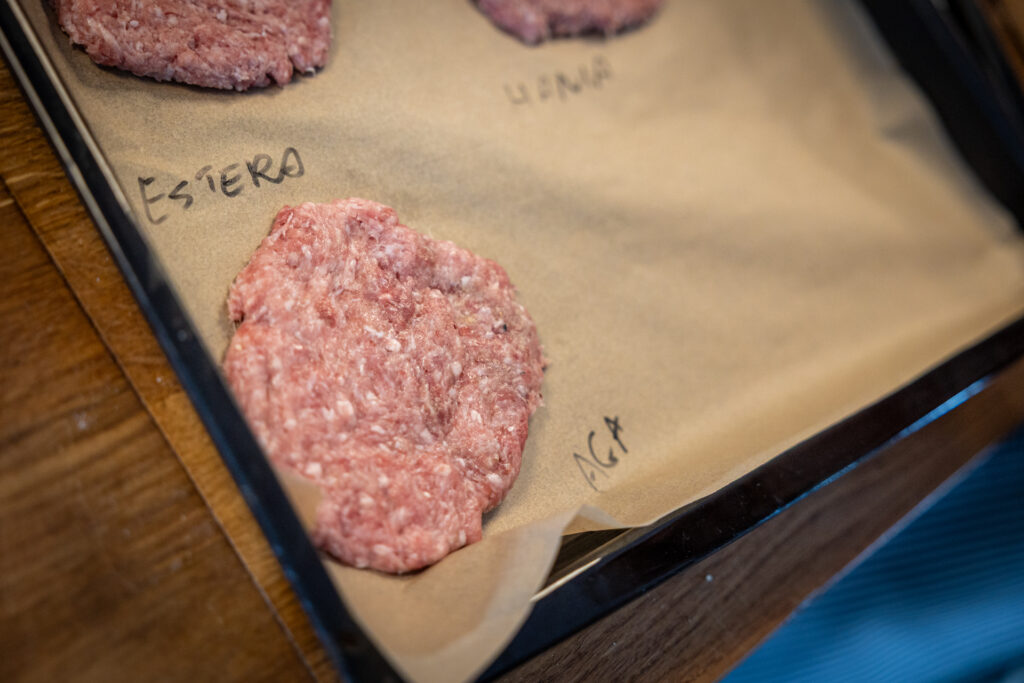 Zdjęcie. Płaski, mielony kotlet leży na papierze do pieczenia na dużej piekarnikowej blasze. Pod nim podpis: Aga. Powyżej dwa inne burgery podpisane Estera i Hania.