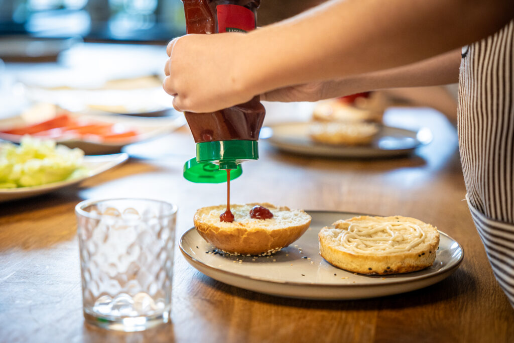 Zdjęcie. Dziecięce dłonie wyciskają ketchup na górną połówkę bułki. Obok na talerzu leży dolna część bułki pokryta majonezem.