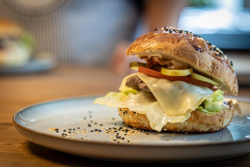 Zdjęcie. Cheesburger na talerzyku. Bułka obsypana jest białymi i kolorowymi ziarenkami. W środku rozciętej bułki sałata, biurger z roztopionym na nim serem, pomidor, ogórek kiszony i prażona cebulka. Pod górną częścią bułki widoczny biały sos. To majonez kielecki.
