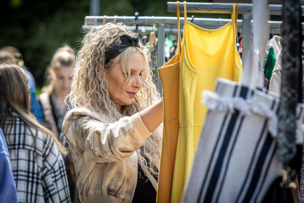 Zdjęcie. Blondynka o pofalowanych włosach z czarną opaską nad czołem ogląda dwie sukienki na ramiączkach, w kolorze miodowym i żółtym.