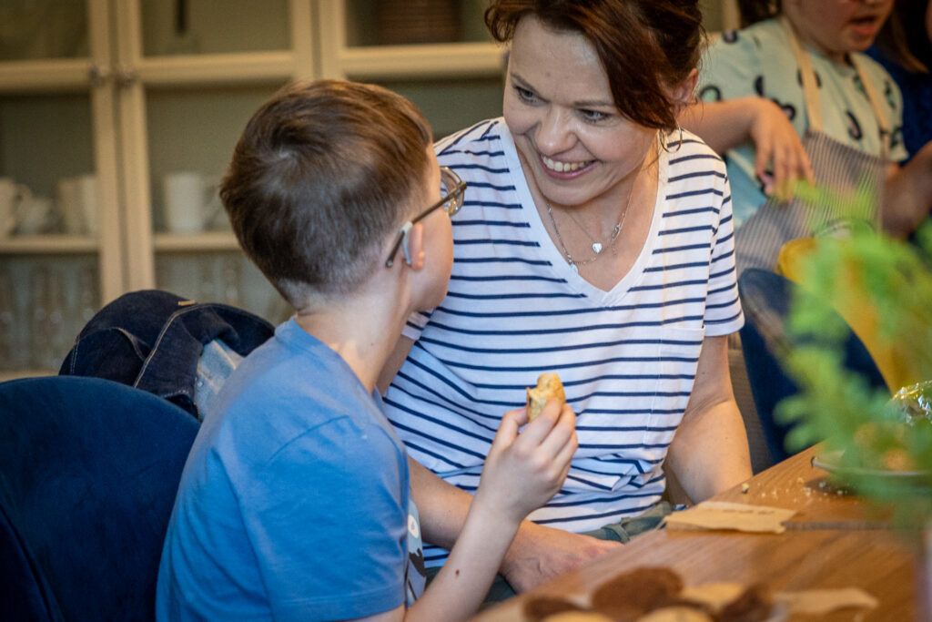 Zdjęcie. Kobieta siedzi obok chłopca. Odwraca się w jego kierunku i bardzo serdecznie się uśmiecha. Obok chłopiec, który patrzy na nią. Ma okulary z grubymi szkłami.
