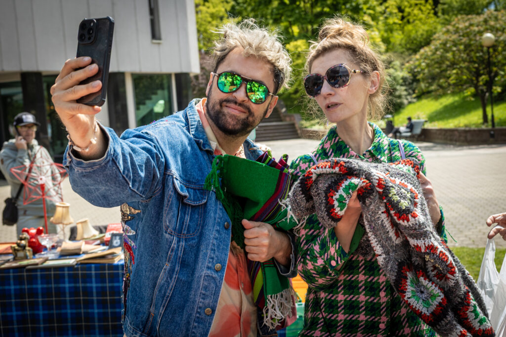 Zdjęcie. Artur Wijata robi selfie sobie i Agacie Buzek. To wysoka szczupła kobieta w płaszczyku w kolorze intensywnej zieleni. Płaszczyk ma deseń: jasnoróżową i czarną kratę.