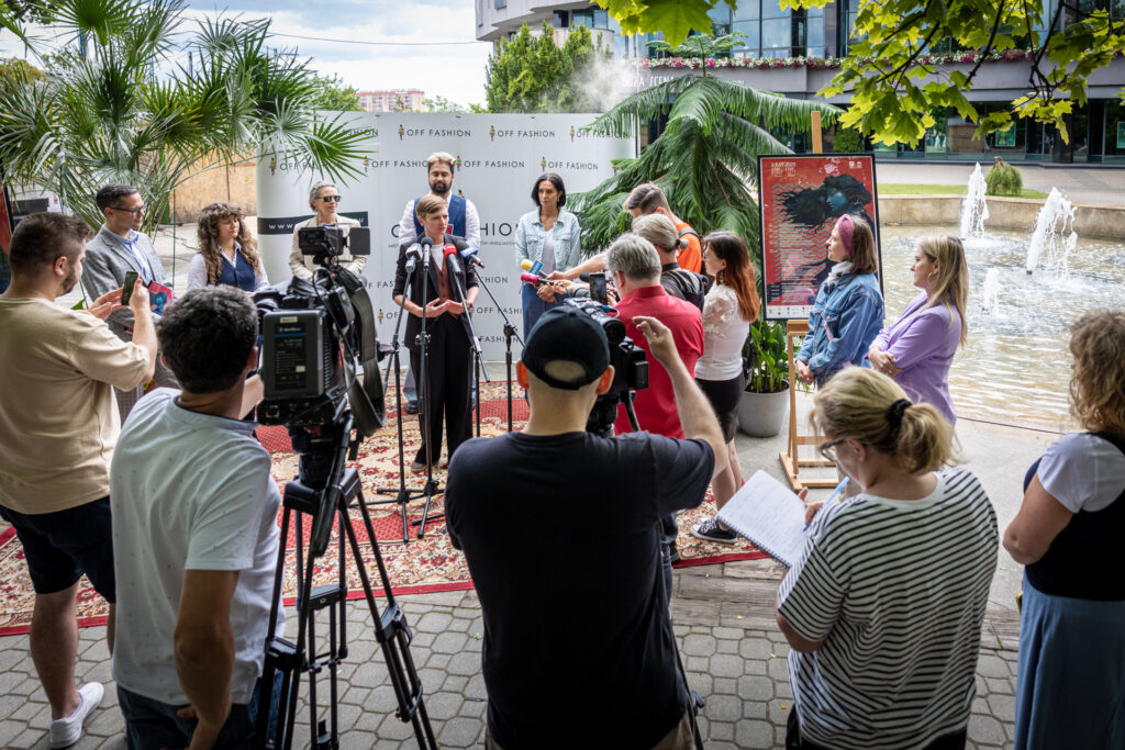 Zdjęcie. Konferencja prasowa poświęcona OFF Fashion. Konferencja odbya się w plenerze, przy fontannie przed KCK. Na kolorowym dywanie stoją osoby uczestnicy konferencji. Przed nimi dziennikarze z kamerami, mikrofonami.