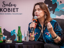 Zdjęcie. Brunetka we wzorzystej kurtce typu bomberka mówi do mikrofonu. To aktorka Renata Dancewicz.